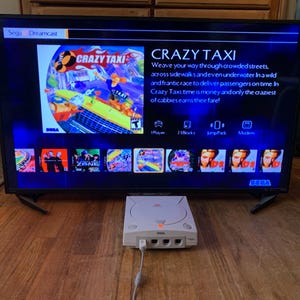 Puede incluir: Una consola de videojuegos Sega Dreamcast blanca con una luz de encendido naranja brillante, conectada a una pantalla de televisión que muestra el menú del juego Crazy Taxi. La pantalla muestra información del juego, otros títulos y el logo de Sega Dreamcast.