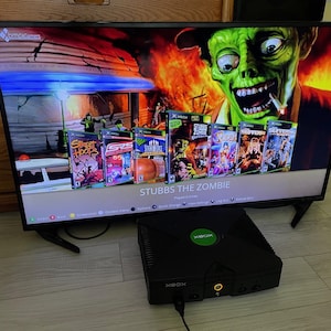 Può includere: Una console di gioco Xbox originale nera è posizionata davanti a uno schermo televisivo che mostra una libreria digitale di giochi Xbox. Lo schermo presenta una grafica prominente di uno zombie verde e un elenco di titoli tra cui Stubbs the Zombie.