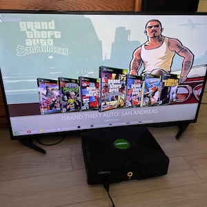 Peut inclure: Une console de jeu Xbox originale noire est posée sur un sol clair devant un écran de télévision affichant le menu du jeu Grand Theft Auto: San Andreas, avec une illustration de personnage et une rangée de diverses jaquettes de jeux Xbox.