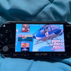 Pode incluir: Uma consola de jogos portátil Sony PSP preta com um ecrã largo que mostra o menu do jogo Grand Theft Auto Vice City Stories, com o texto Tuesday, 14. November 2023 1:35:25 64% (4:17) MODEST SYSTEMS PSP visível no topo do ecrã.