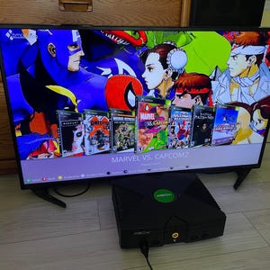 以下が含まれることがあります： Marvel vs. Capcom 2のメニュー画面が表示されたテレビの前に置かれた黒いオリジナルのXboxゲーム機。画面にはManhunt、Getting Up、Marvel vs. Capcom 2などの様々なゲームのパッケージが表示されています。