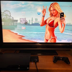 May include: A black Sony television screen displaying a Grand Theft Auto V game menu with a beach scene and a woman in a red bikini, positioned above a black PlayStation 3 console, a black game controller, and a small digital clock showing 59.