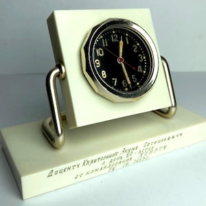 Könnte beinhalten: Eine Tischuhr mit schwarzem, rundem Zifferblatt und goldfarbener Lünette auf einem quadratischen, cremeweißen Sockel. Die Uhr wird von zwei goldfarbenen Metallarmen gehalten, die auf einem rechteckigen, cremeweißen Standfuß mit kyrillischer Gravur sitzen.