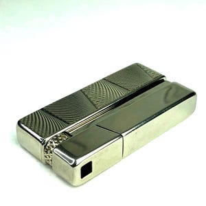 May include: A rectangular silver-toned metal lighter featuring a polished smooth finish on one side and a textured, cross-hatched pattern on the other. The device includes a small, knurled circular ignition wheel and a square opening for the flame.
