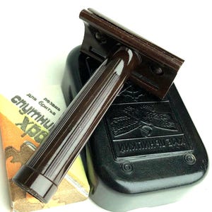 May include: A dark brown safety razor with a ribbed handle resting on a black rectangular case. Beside the case is a small rectangular paper box with yellow and white colouring, featuring Cyrillic text including the word "Спутник".