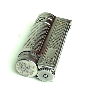 May include: A silver-toned metal lighter with a textured, cross-hatched pattern on its cylindrical body. The base features the engraved text "Oreo Pat Austria". It includes a small circular dial on the side and a smooth rectangular panel.