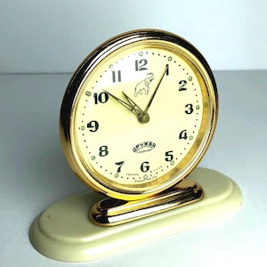 Può includere: Un orologio da tavolo analogico rotondo con cornice in metallo dorato e quadrante color crema. Il quadrante presenta numeri arabi neri, un'immagine centrale di un elefante e testo in cirillico. È montato su una base ovale in plastica bianco sporco.