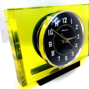 Pode incluir: Um relógio de mesa retangular com uma moldura de bloco de acrílico amarelo neon translúcido. O mostrador circular é preto com algarismos arábicos brancos e ponteiros prateados, envolto numa borda de metal prateado polido sobre uma base preta.
