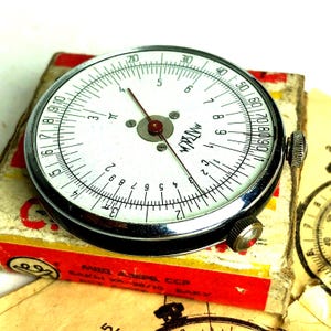 Peut inclure: Une règle à calcul mécanique circulaire avec un cadran blanc, des graduations numériques noires et une fine aiguille indicatrice rouge. L'appareil présente un bord argenté métallique et deux petits boutons de réglage moletés sur le côté.