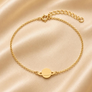 Könnte beinhalten: Ein goldfarbenes Armband mit einem zentralen Anhänger in Form eines Planeten mit einem Ring in der Mitte. Das Armband besteht aus einer feinen Kette mit einem runden Federringverschluss und einer verstellbaren Verlängerungskette am Ende.