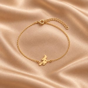 Op de afbeelding: Een goudkleurige metalen kettingarmband met een centrale bedel in de vorm van een silhouet van een heks op een bezemsteel. De armband is voorzien van een fijne schakelketting en een ronde sluiting aan het uiteinde voor een veilige bevestiging.