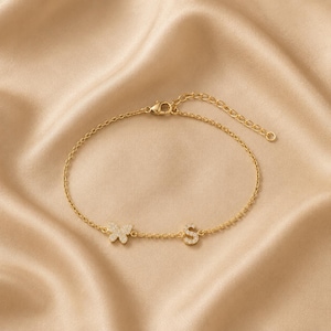 Puede incluir: Una pulsera de cadena en tono dorado con un pequeño dije de mariposa y un dije con la letra S, ambos incrustados con piedras pavé transparentes. La pulsera incluye una delicada cadena de eslabones y un cierre de mosquetón con extensión.