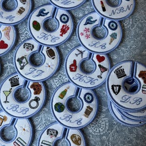 May include: A collection of round, white fabric clothing rack dividers with light blue stitched borders. Each divider features unique, colorful embroidered icons such as hearts, sports gear, food, and animals, along with custom embroidered initials.
