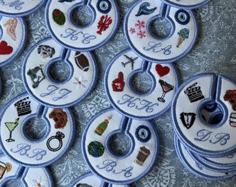 May include: A collection of round, white fabric clothing rack dividers with light blue stitched borders. Each divider features unique, colorful embroidered icons such as hearts, sports gear, food, and animals, along with custom embroidered initials.