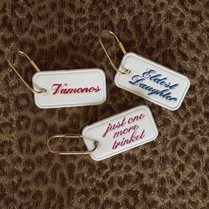 May include: Three rectangular white fabric keychains with gold-toned metal carabiner clips. Each keychain features embroidered text: one in red reading Vamonos, one in blue reading Eldest Daughter, and one in dark pink reading just one more trinket.
