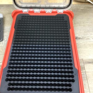 May include: A rectangular red storage case with an open clear lid and a black foam insert featuring a grid of circular indentations. The interior foam is designed with rows of small, uniform holes for organizing items within the hard plastic shell.