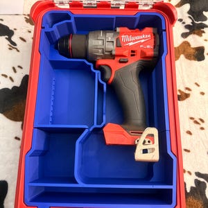 May include: A red Milwaukee Fuel cordless drill with a black grip is stored inside a custom blue plastic insert within a red hard-shell carrying case. The drill features a metal belt clip and clear markings on the chuck. The case has a transparent lid.