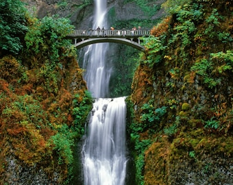 May include: A scenic photograph of a waterfall cascading down a rocky cliff face. A stone bridge arches over the falls, with several people visible on the bridge. Lush green and orange foliage surrounds the waterfall.