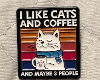 May include: A rectangular enamel pin featuring a white cat wearing a blue scarf and holding a coffee mug. The background has horizontal stripes in orange, red, and purple. The text reads I LIKE CATS AND COFFEE AND MAYBE 3 PEOPLE in white.