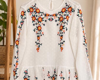 Könnte beinhalten: Eine weiße Langarmbluse aus strukturiertem Swiss-Dot-Stoff mit einem gerüschten Saum. Das Kleidungsstück ist mit farbenfrohen Blumenstickereien in Rot-, Orange-, Gelb- und Grüntönen auf der Brust, den Ärmeln und am unteren Saum verziert.