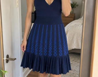 Könnte beinhalten: Ein ärmelloses Strickkleid in Marineblau mit tiefem V-Ausschnitt, breitem Rippstrickbund und einem ausgestellten Rock mit vertikalen blauen geometrischen Mustern. Der Rock endet in einem einfarbigen, marineblauen Plissee-Saum.