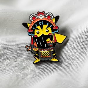 May include: A metal enamel pin featuring a yellow character with pointed ears dressed in traditional Chinese opera attire. The character wears an ornate red, black, and white headdress with a small sphere, a black beard, and a black and gold patterned robe.