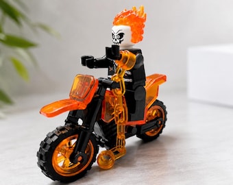 Könnte beinhalten: Eine Kunststoff-Minifigur mit weißem Totenkopf-Kopf und durchscheinendem orangefarbenem Flammenhaar, die einen schwarzen Oberkörper mit weißen Grafikdetails trägt. Die Figur sitzt auf einem schwarzen und orangefarbenen Spielzeugmotorrad.