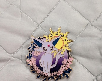May include: A circular enamel pin featuring a light purple feline creature with large ears and a red forehead gem. The design includes a yellow sun, a dark purple background, and a border of pink flowers and dark blue leaves with gold metal outlines.