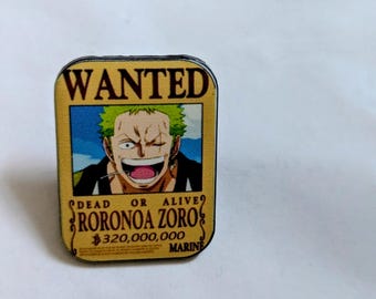 Könnte beinhalten: Ein rechteckiger Metall-Pin mit einem gelben Steckbrief-Design. Der Text lautet WANTED, DEAD OR ALIVE, RORONOA ZORO, 320.000.000 und MARINE. In der Mitte befindet sich eine grafische Illustration einer Figur mit grünen Haaren.