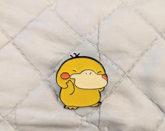 May include: A yellow enamel pin shaped like a cartoon duck-like creature with a cream-colored beak, small black eyes, and round orange cheeks. The character has a rounded body, two small feet, and three tiny black tufts of hair on its head.