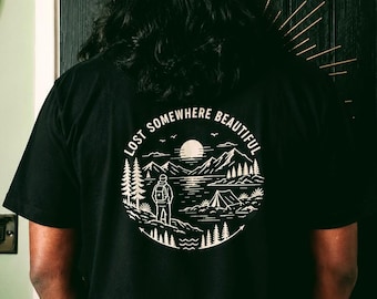 Könnte beinhalten: Ein schwarzes T-Shirt mit einer kreisförmigen weißen Grafik auf der Rückseite. Das Design zeigt eine Person mit Rucksack an einem See, Bergen, Tannen und einem Zelt unter einer Sonne. Der Text "LOST SOMEWHERE BEAUTIFUL" steht darüber.