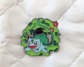 May include: A metal enamel pin featuring a light blue creature with a large green bulb on its back, positioned within a circular frame of green vines and small red flowers. The design includes black outlines and a glossy finish on the metal surface.