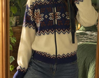 Könnte beinhalten: Eine cremefarbene Strickjacke mit Reißverschluss, dunkelblauer Passe und Saum sowie einem sich wiederholenden geometrischen Schneeflockenmuster in Weiß, Braun und Blau. Das Kleidungsstück hat lange Ärmel mit Rippbündchen und einen Reißverschluss.