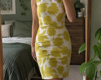 Könnte beinhalten: Ein ärmelloses, knielanges Kleid mit weißem Grund und einem sich wiederholenden Muster aus gelben Sonnenblumen. Das Kleidungsstück hat einen Rundhalsausschnitt, ein eng anliegendes Oberteil und einen geraden Rock, der knapp über dem Knie endet.