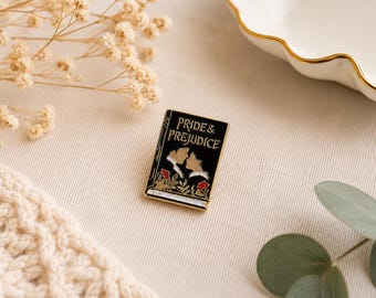 May include: A rectangular enamel pin featuring a black book cover with the gold text Pride & Prejudice. The design includes a silhouette of two figures facing each other, accented by small red flowers and green leaves at the bottom edge.
