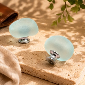 May include: Two cabinet knobs featuring smooth, rounded, frosted light blue glass tops attached to polished silver-tone metal bases. The glass has a soft, matte texture, and the metal hardware includes a circular mounting plate at the bottom.