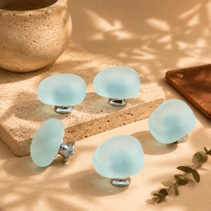 May include: A set of four cabinet knobs featuring smooth, rounded, frosted light blue glass tops attached to polished silver metal bases. Each knob has a pebble-like shape with a matte, translucent finish, providing a consistent look.