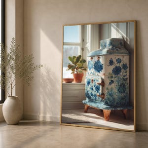 May include: A large rectangular wall mirror with a thin gold frame reflecting a room interior. The reflection shows a tiled stove decorated with blue floral patterns, a potted plant on a windowsill, and a light-colored floor with a rug.