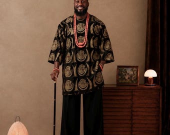 Könnte beinhalten: Eine schwarze Tunika mit goldenem Kreismuster, kombiniert mit einer schwarzen Hose, einer passenden gemusterten Kopfbedeckung und einer mehrreihigen Korallenperlenkette. Das Ensemble umfasst einen schwarzen Gehstock mit Goldgriff und ein Korallenarmband.