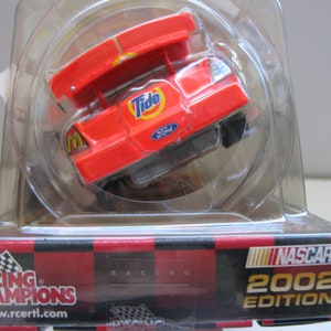 Może przedstawiać: Czerwono-biało-niebieski model samochodu NASCAR Racing Champions z 2002 roku z logo Tide i Ford. Samoch&oacute;d znajduje się w przezroczystym plastikowym pudełku.