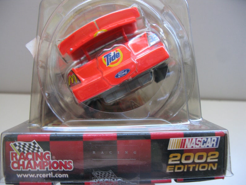 Może przedstawiać: Czerwono-biało-niebieski model samochodu NASCAR Racing Champions z 2002 roku z logo Tide i Ford. Samoch&oacute;d znajduje się w przezroczystym plastikowym pudełku.