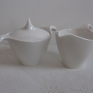 May include: A white ceramic sugar bowl and creamer set with a unique, modern design. The sugar bowl has a lid with a pointed top and a curved handle. The creamer has a curved handle and a wide, open top.