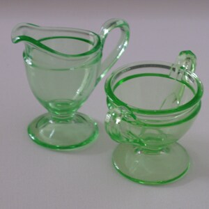May include: A vintage green glass creamer and sugar bowl set. The creamer has a handle and a rounded base. The sugar bowl has a lid and two handles.