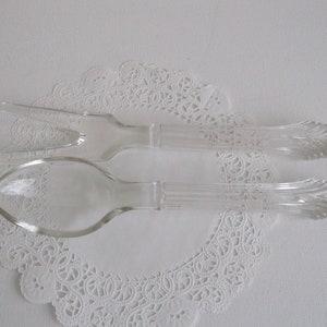 May include: A set of two clear glass serving utensils, a spoon and a fork, with decorative detailing on the handles. The utensils are on a white doily.
