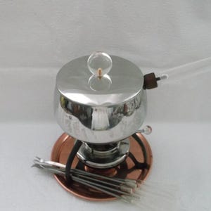May include: A stainless steel fondue pot with a copper-colored base and a clear glass handle. The pot has a lid with a clear knob and is accompanied by several fondue forks. The set is designed for serving melted cheese or chocolate.