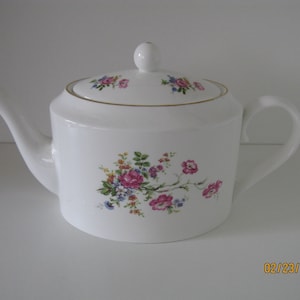 May include: White porcelain teapot with a lid, spout, and handle. The teapot is decorated with a floral pattern featuring pink, blue, and yellow flowers. A gold trim accents the rim and lid. The teapot is designed for serving tea.