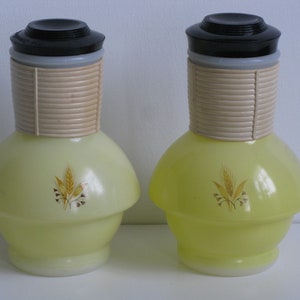 May include: Two yellow glass pitchers with black lids and a beige band around the neck. Each pitcher has a wheat design on the front.