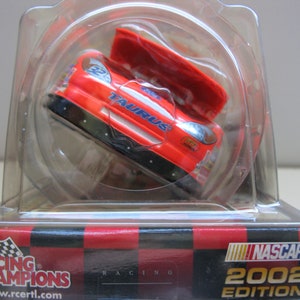Może przedstawiać: Czerwono-biały model samochodu NASCAR Racing Champions z edycji 2002 w przezroczystej plastikowej gablocie. Samoch&oacute;d to Ford Taurus z otwartym kapturem.