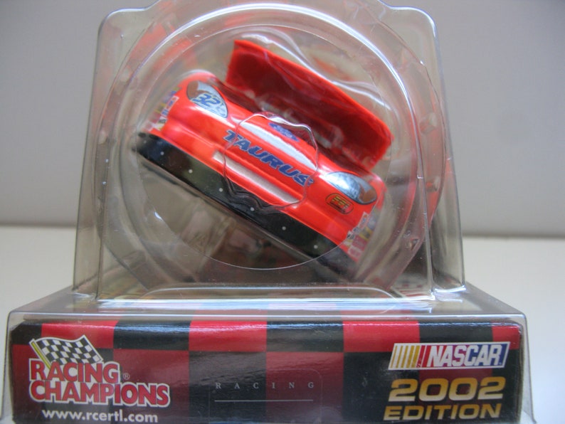 Może przedstawiać: Czerwono-biały model samochodu NASCAR Racing Champions z edycji 2002 w przezroczystej plastikowej gablocie. Samoch&oacute;d to Ford Taurus z otwartym kapturem.