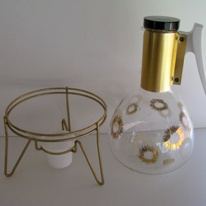 May include: A gold metal stand with a round top and three legs. A glass carafe with a gold lid and a white handle. The carafe has a gold floral pattern.